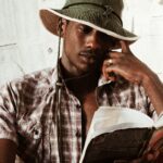 Young African American male in cricket hat and open shirt reading book while sitting near wall with papers with hand on cheek